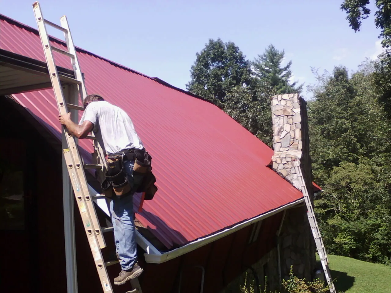 Roofer on a ladder beside a red metal roof and stone chimney — Soffit & Fascia Repair by Roofing Brothers in Cropseyville